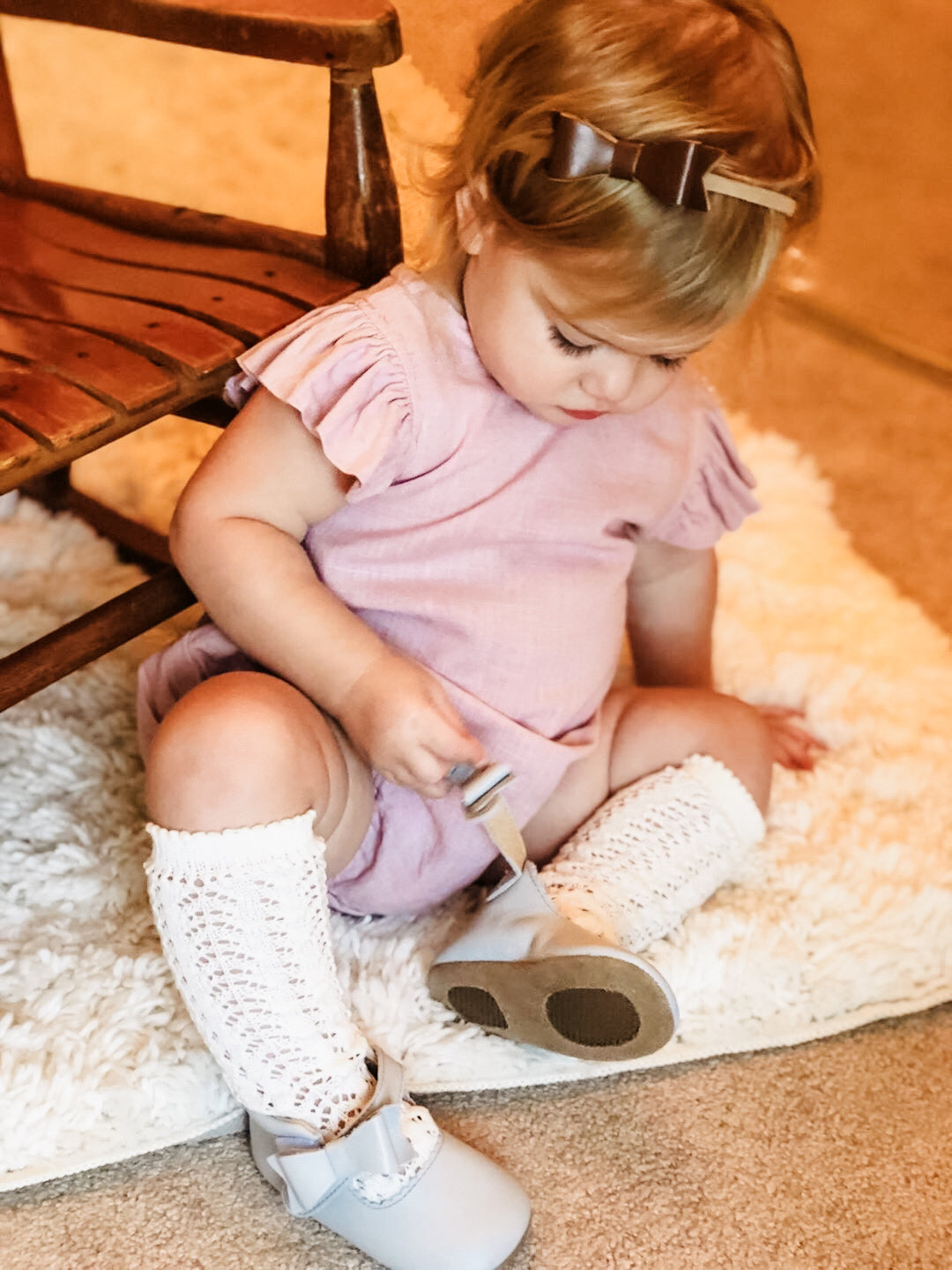 A baby girl in a ruffled lavender romper and white knit knee‑high socks sits on a soft rug, adjusting her light gray Harper & Hedgie bow shoes — blending vintage‑inspired charm with flexible, supportive design for growing feet.