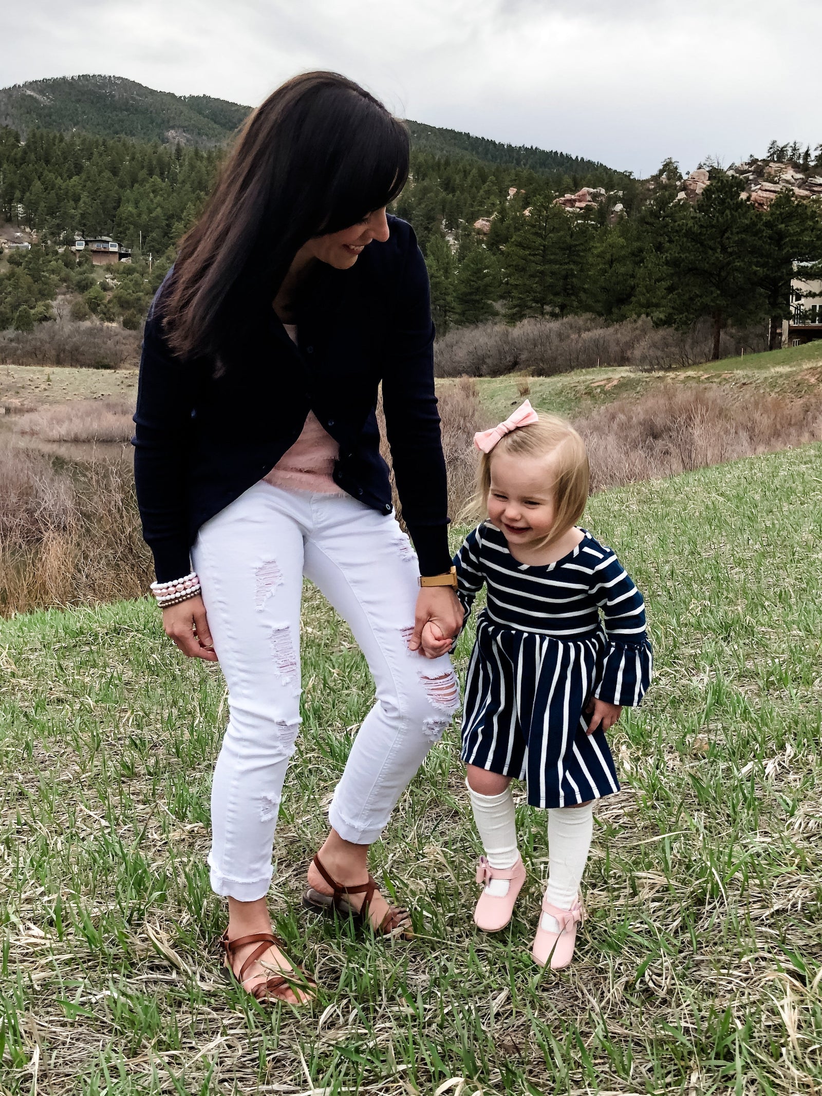 Adult and child holding hands on grass with scenic hills in the background. Child wears striped dress and pink shoes. Peaceful outdoor moment.