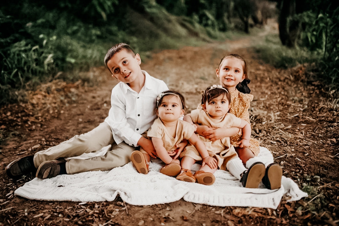 Four young children sitting on a white blanket along a dirt path surrounded by greenery, with toddlers in matching beige outfits and older siblings in classic fall clothing, captured in soft natural light.