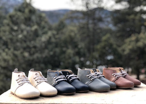 “Four pairs of baby shoes in white, cream, navy, and brown, arranged on a stone surface with a blurred mountain and pine tree backdrop—evoking handcrafted quality, nature, and early childhood warmth.