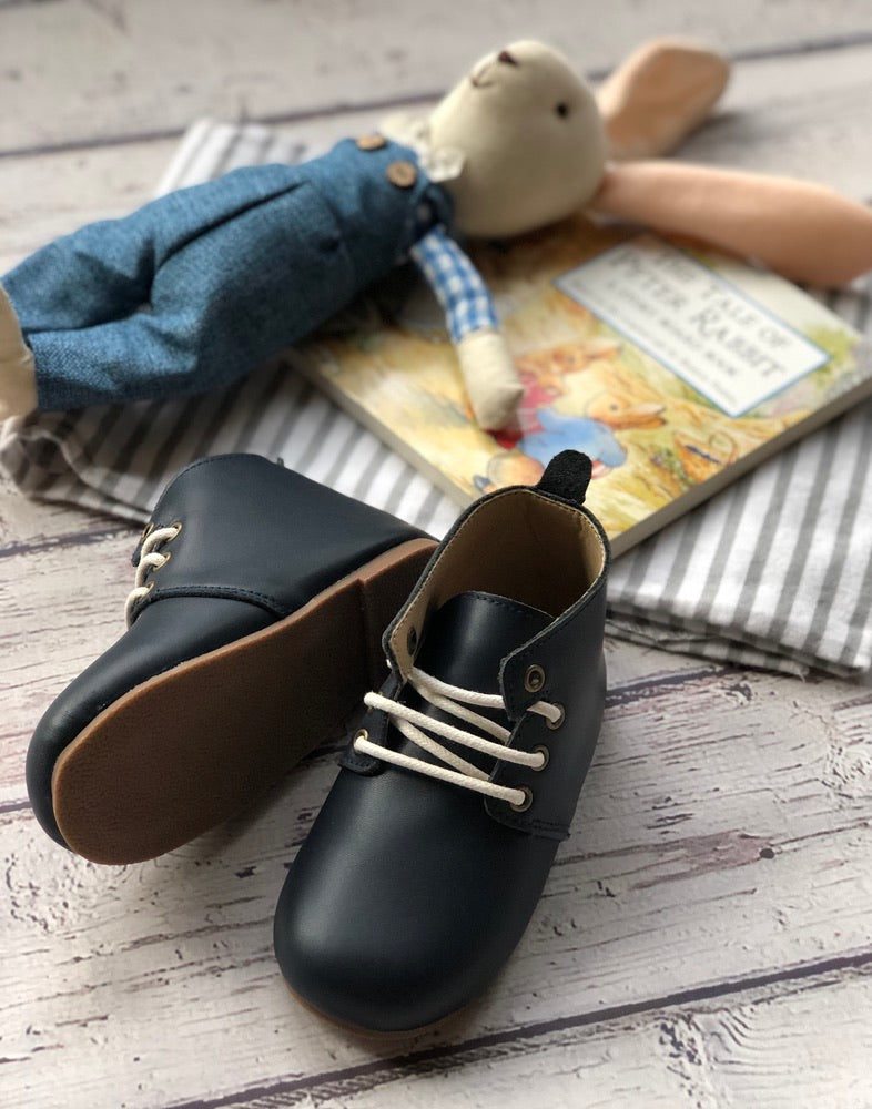 leather baby Oxford shoes with white laces styled on wooden surface beside cloth rabbit doll in blue overalls and “The Tale of Peter Rabbit” book.