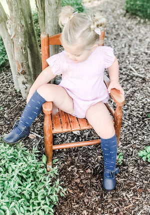 Young child in pink outfit and navy knee‑high socks wearing navy Mary Janes, seated on wooden chair in a garden setting.