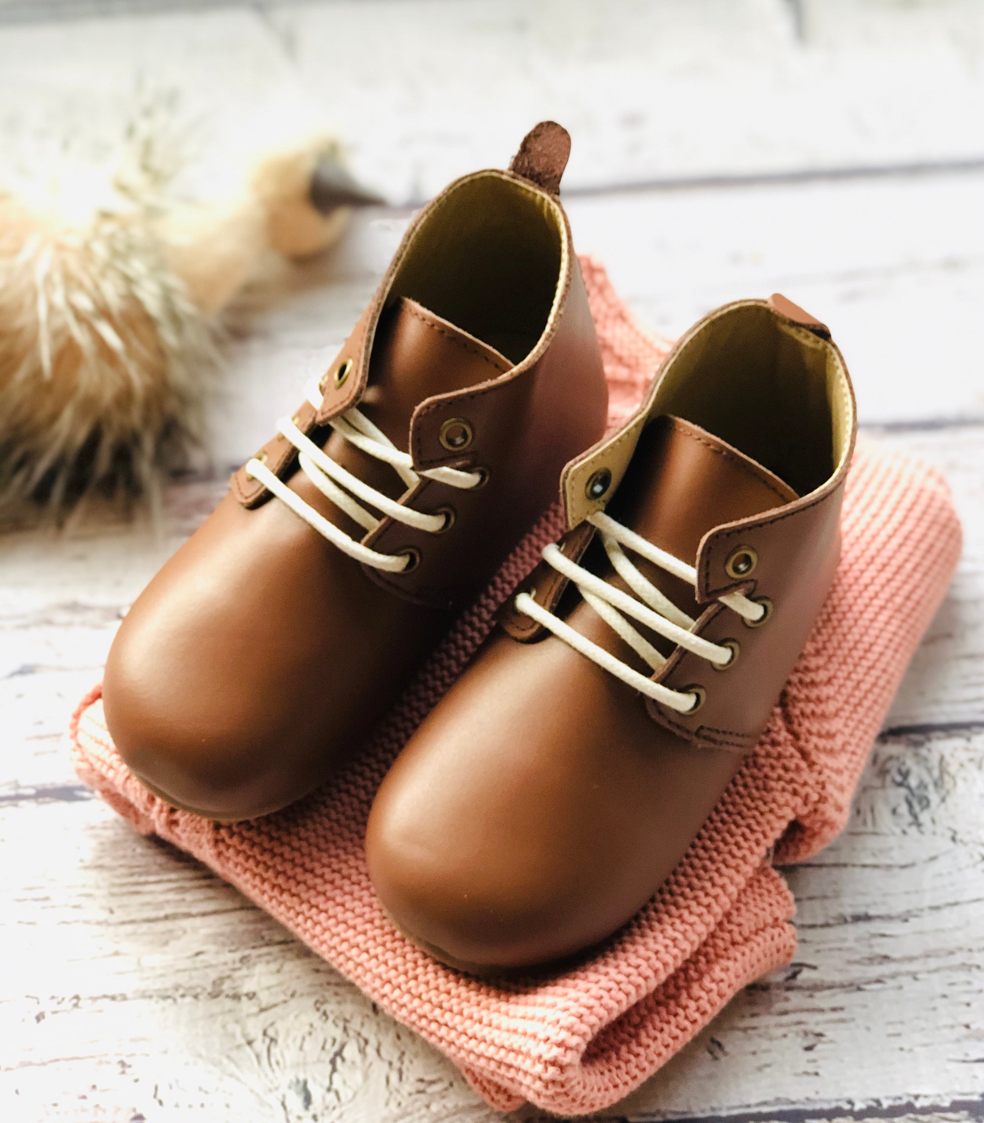 Classic brown leather baby shoes with white laces, styled on folded pink knitted garment for a cozy, rustic nursery aesthetic.