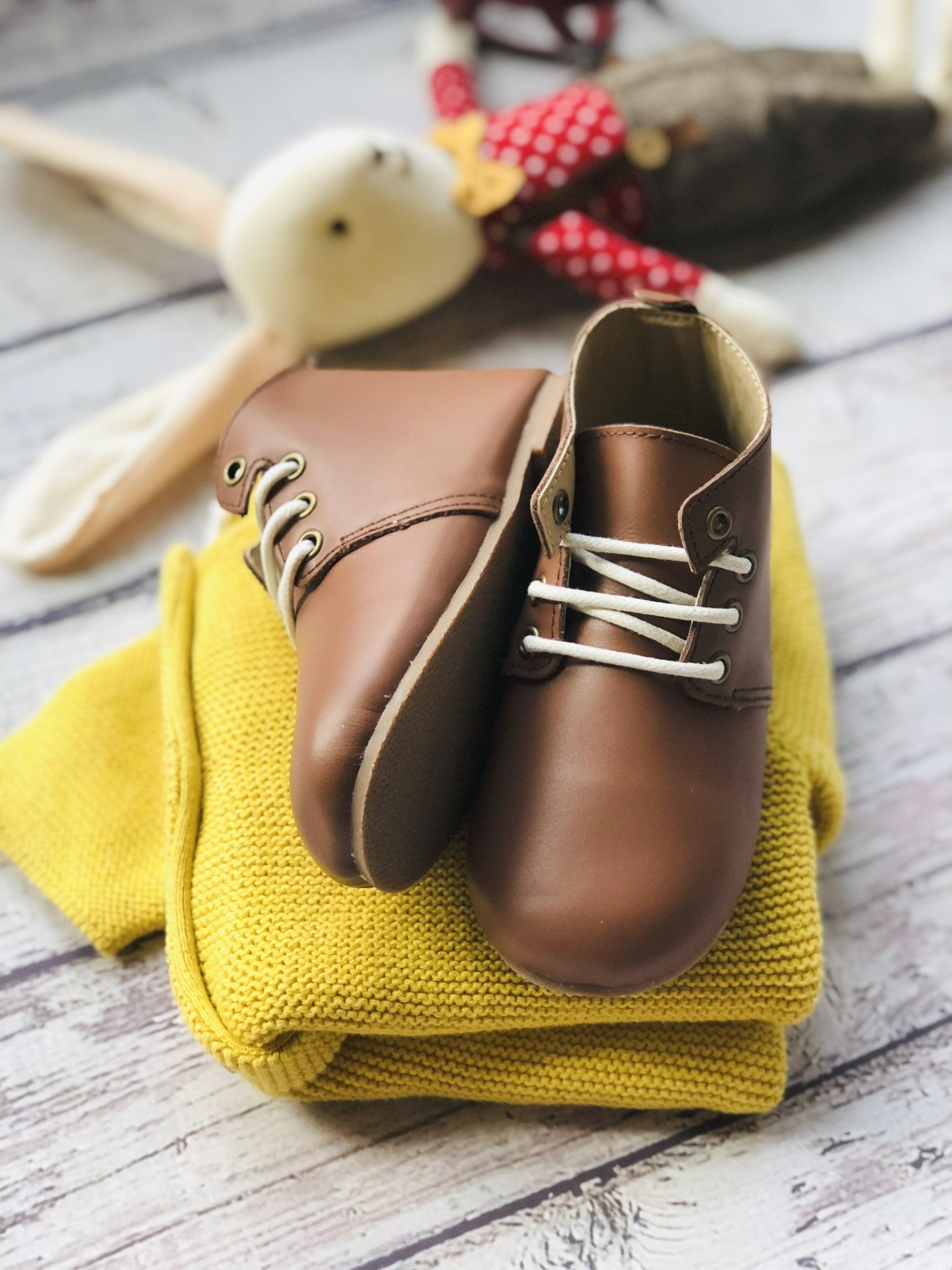 Brown leather children’s shoes with white laces, displayed on folded yellow knit sweater beside plush rabbit toy in red polka‑dot outfit.