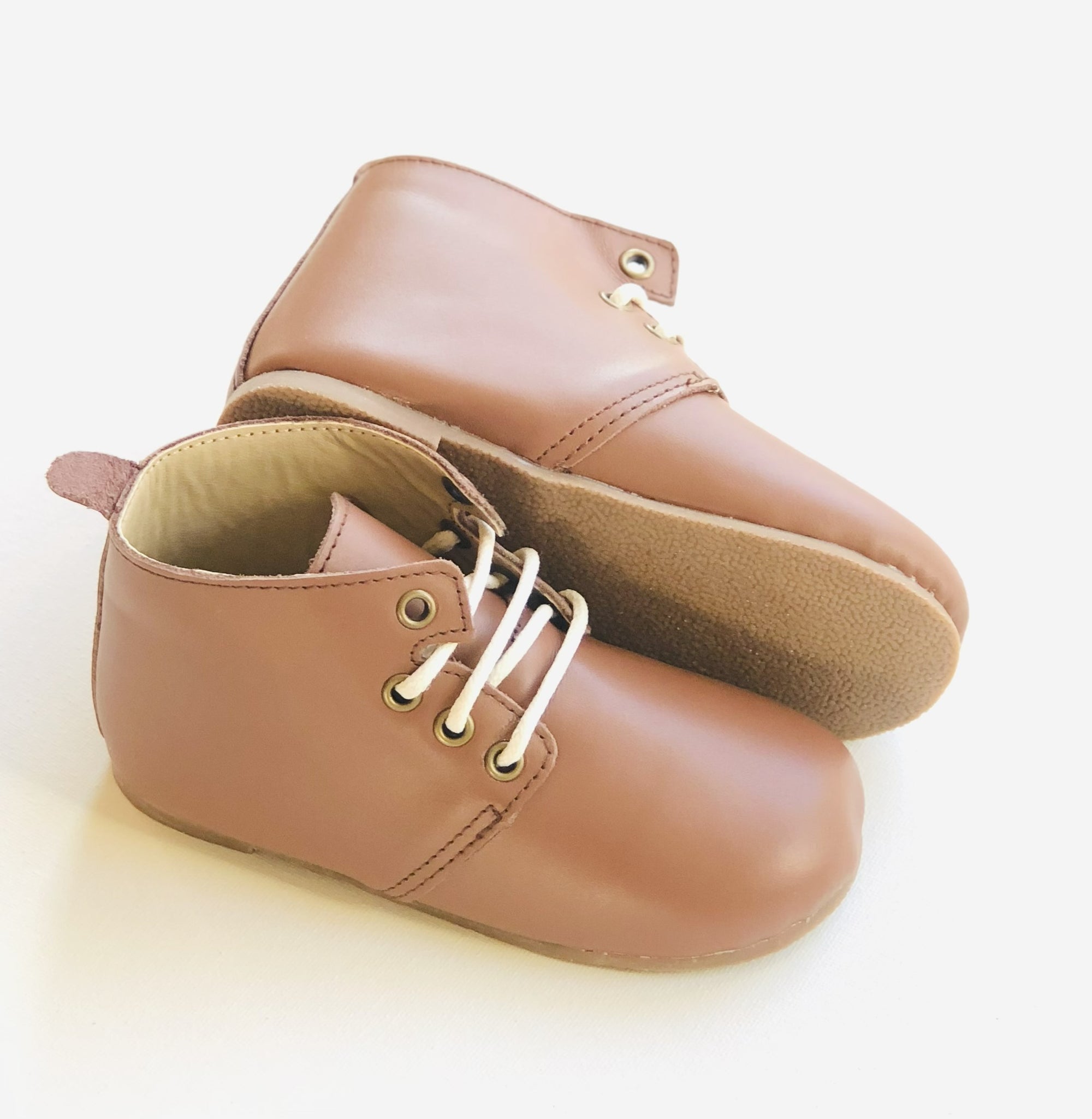Brown leather baby shoes with white laces, soft rubber sole, and metal eyelets — one upright and one on its side to show tread detail.
