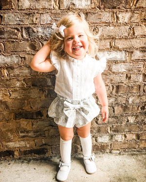 Smiling toddler in white ruffled top, bow-tied shorts, and white shoes, standing against a rustic brick wall.