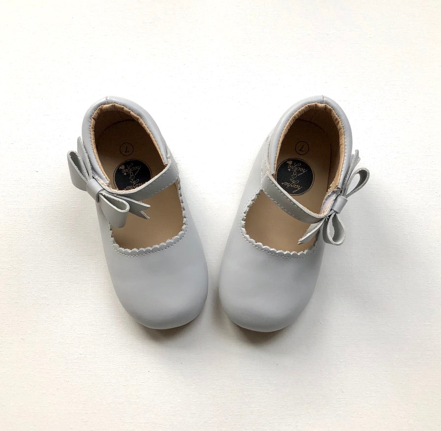 Light gray Mary Jane baby shoes with scalloped trim and decorative bows, styled for infants or toddlers