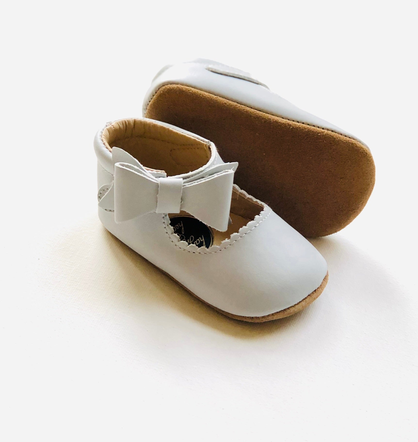 Classic white leather baby Mary Jane shoes with scalloped edges, front bows, and brown sole, one upright and one on its side to show detail.