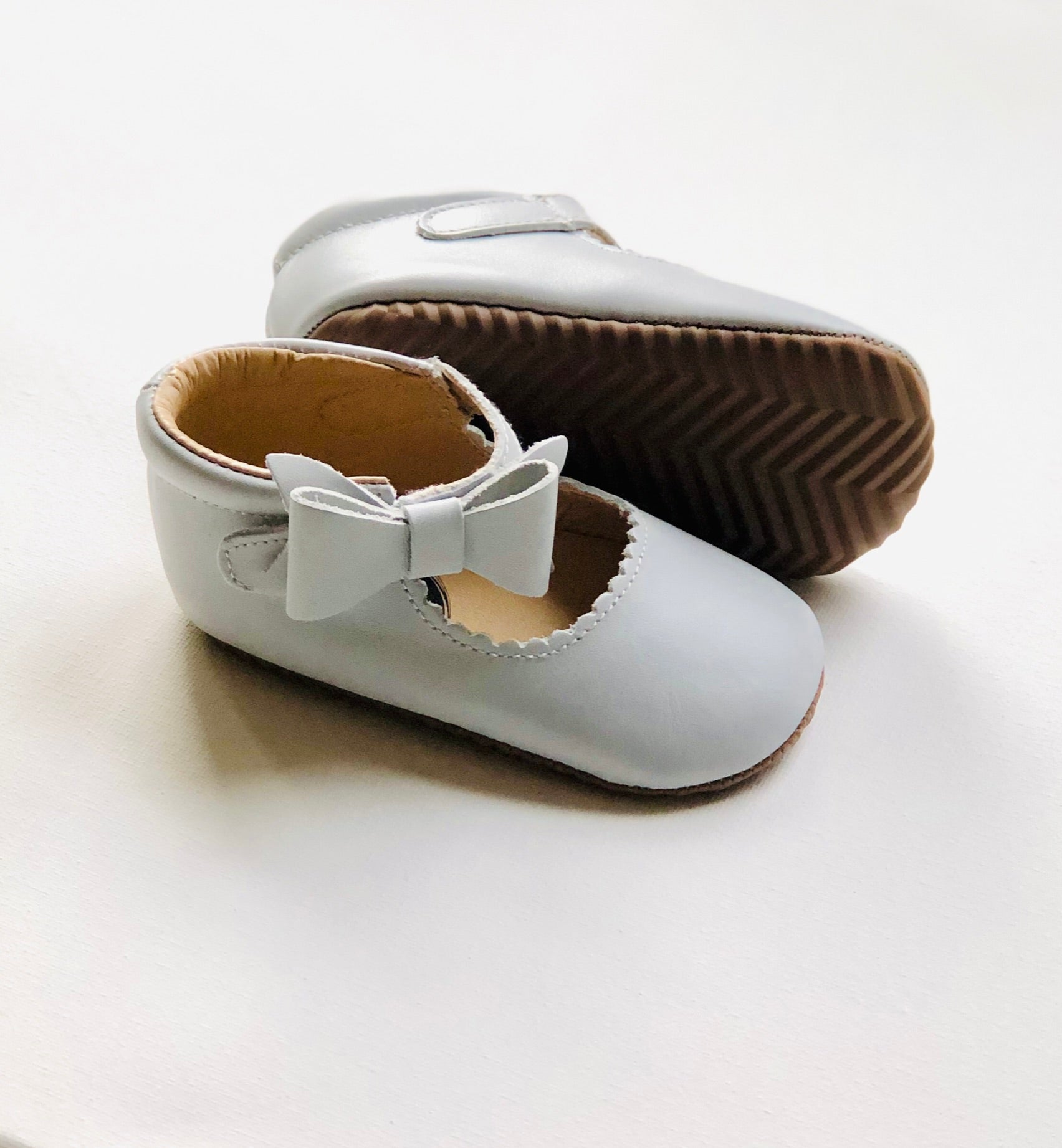 Light gray leather baby Mary Jane shoes with scalloped trim, front bows, and zigzag tread sole, one upright and one on its side for full view.