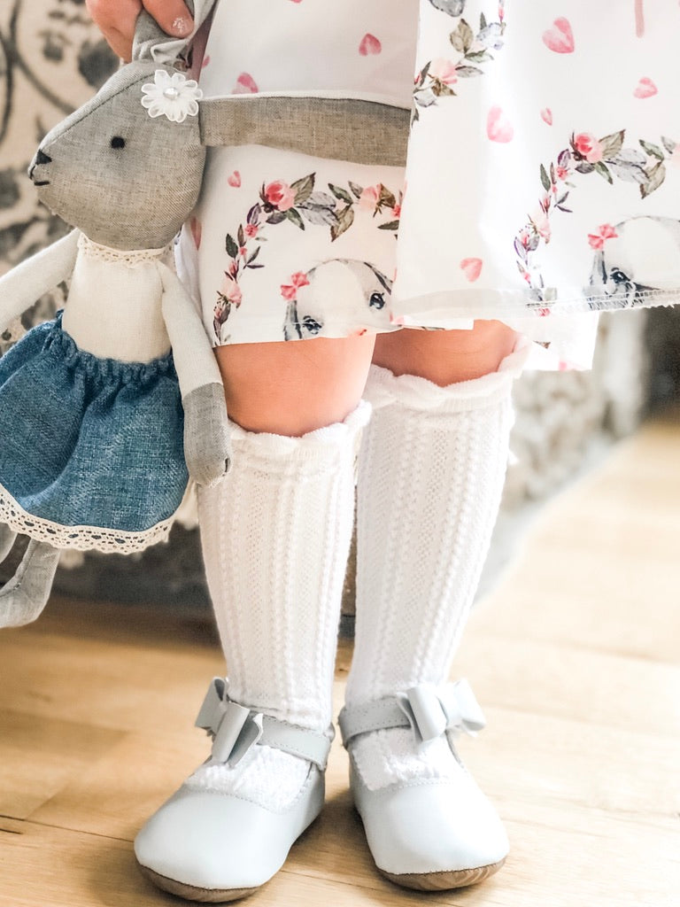 Toddler in floral dress and lace‑trim socks wearing light gray Mary Jane shoes, holding plush bunny in blue skirt, in a soft pastel indoor setting.