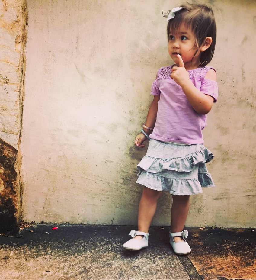 Young child in lace‑shoulder top and ruffled skirt wearing white ankle‑strap shoes, standing against rustic textured wall for a charming, candid moment.