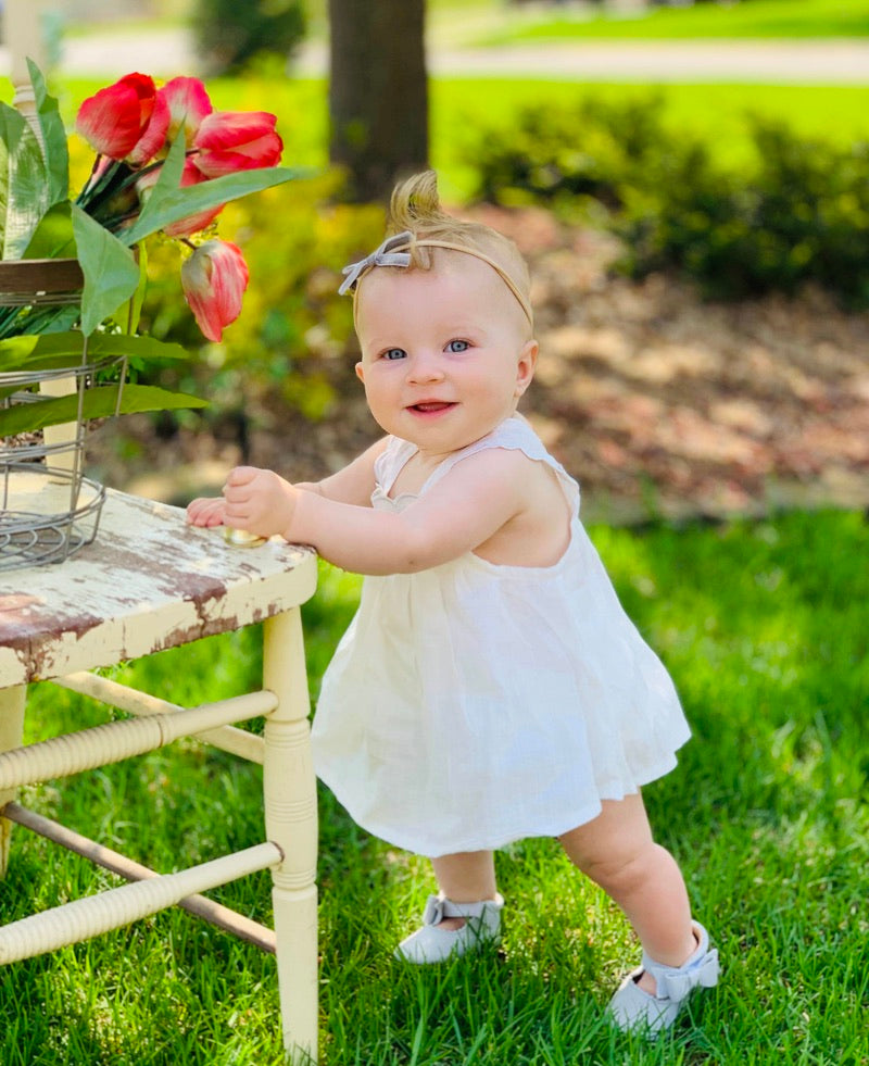 Toddler in white dress and bow headband wearing white shoes, holding rustic chair with basket of red tulips in a sunny garden setting.