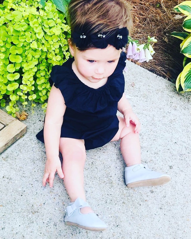 Toddler in black ruffled outfit and headband wearing light gray strap shoes, sitting on concrete near green plants and purple flowers in a garden scene.
