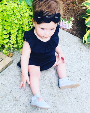 Toddler in black ruffled outfit and headband wearing light gray strap shoes, sitting on concrete near green plants and purple flowers in a garden scene.