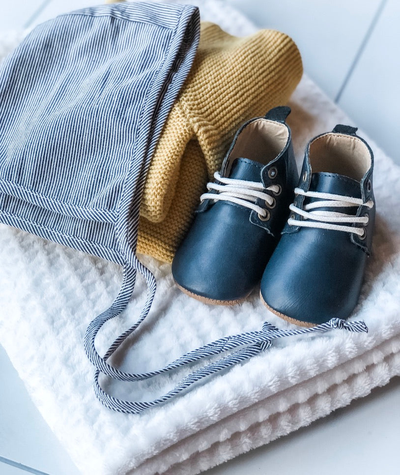 Flat lay of navy baby shoes with white laces, striped bonnet, and mustard knitwear – cozy and curated infant fashion