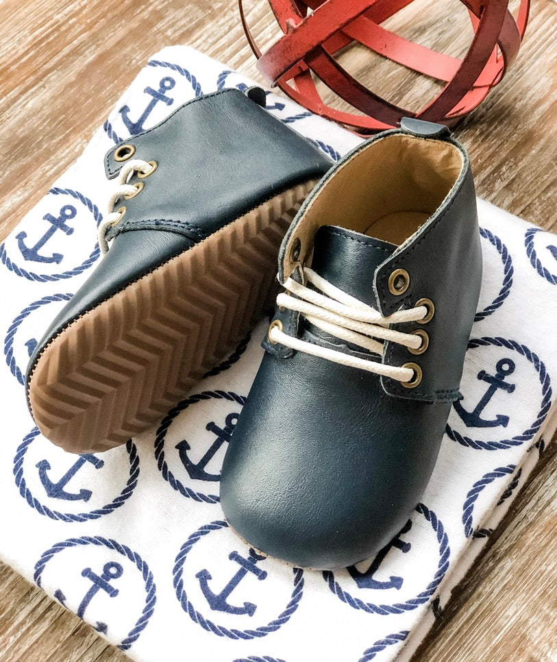 Navy baby shoes with white laces and gold eyelets on anchor-patterned cloth – nautical-inspired toddler footwear with zigzag sole