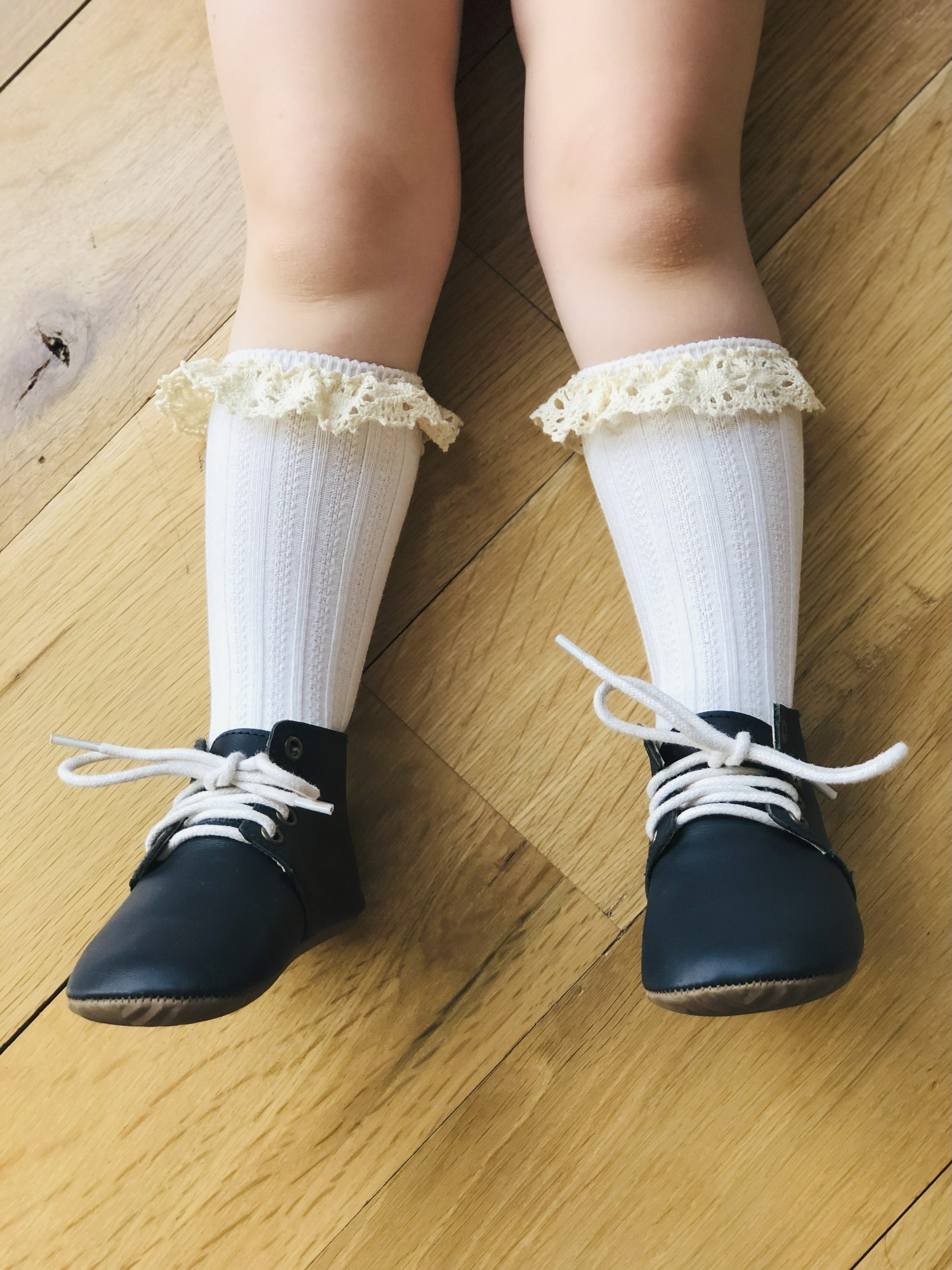 Toddler wearing black leather shoes with white laces and lace-trimmed socks – vintage-inspired children’s fashion on wooden floor