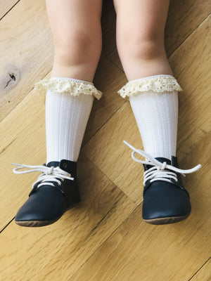 Toddler wearing black leather shoes with white laces and lace-trimmed socks – vintage-inspired children’s fashion on wooden floor