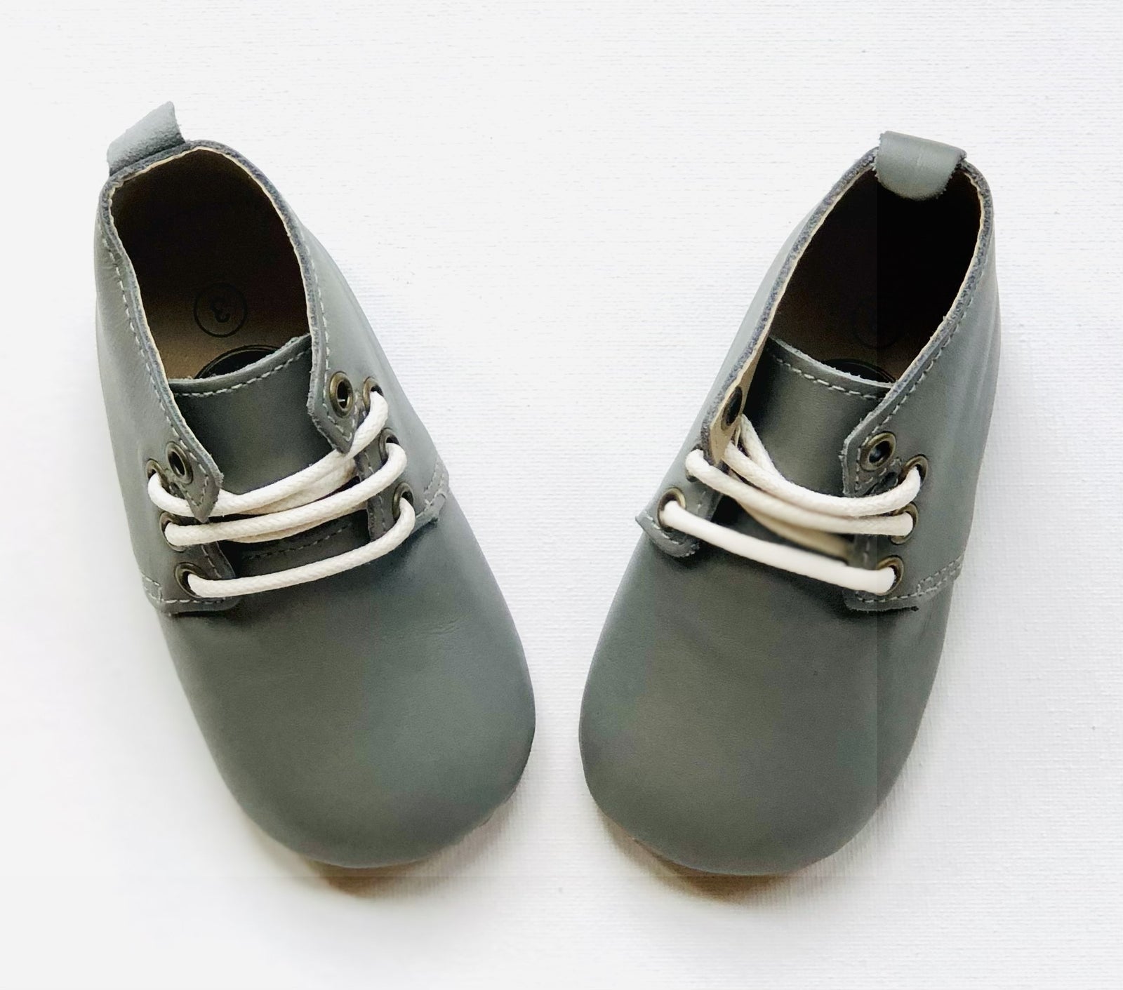 Small gray leather baby shoes with white laces and rounded toes, placed side by side on white background for product clarity.