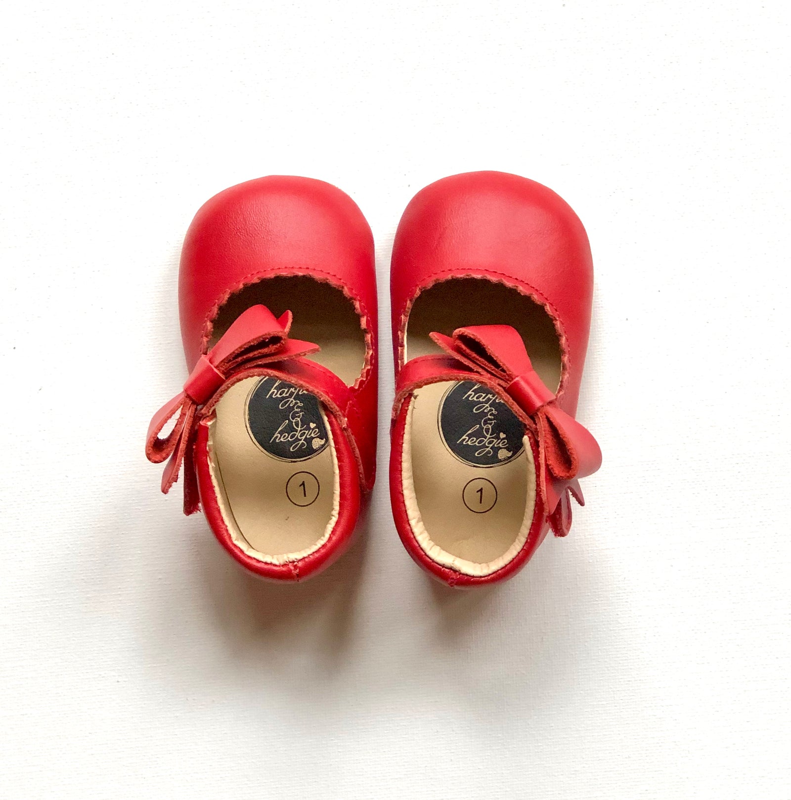 Red leather baby Mary Janes with scalloped trim and side bows, featuring Harper & Hedgie branding and size label.