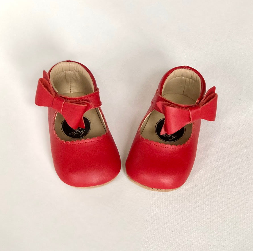 Red baby shoes with front bows and rounded toes, styled on white surface with soft beige interior lining.