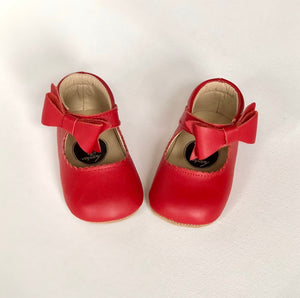 Red baby shoes with front bows and rounded toes, styled on white surface with soft beige interior lining.