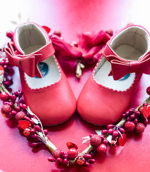 Valentine’s-themed flat lay of red baby shoes with bows, surrounded by heart-shaped berry and glitter decor on pink background.