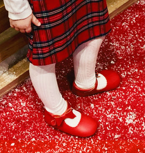 Toddler in red plaid dress, white tights, and red bow shoes standing on festive surface with artificial snow.