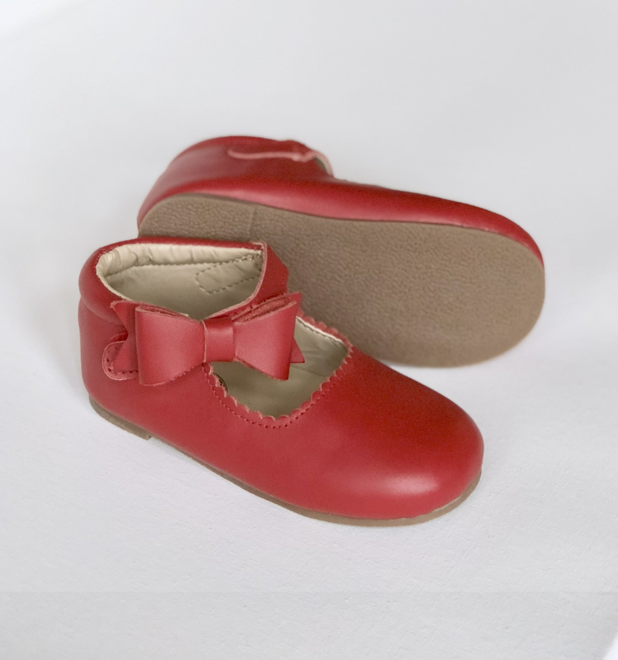 Red baby shoes with scalloped edges, side bow, and soft beige sole, shown upright and side view for detail.