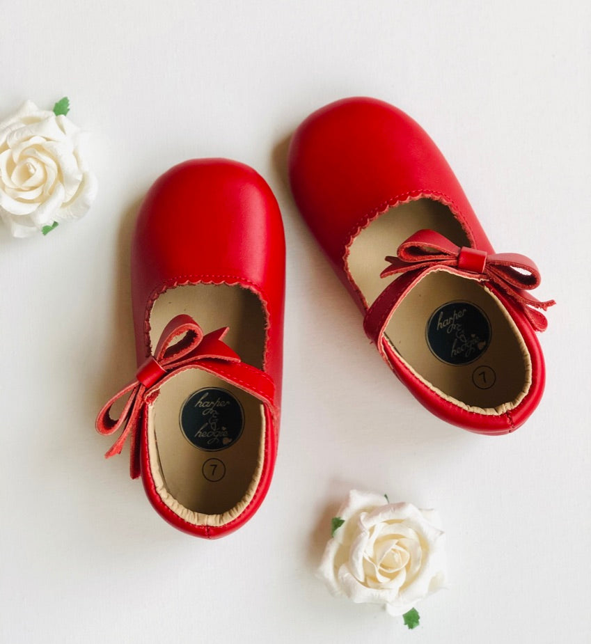 Red leather baby shoes with scalloped trim and bow detail, styled with white artificial roses on white background.