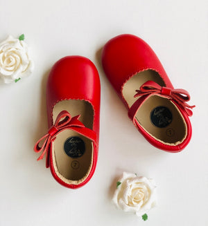 Red leather baby shoes with scalloped trim and bow detail, styled with white artificial roses on white background.