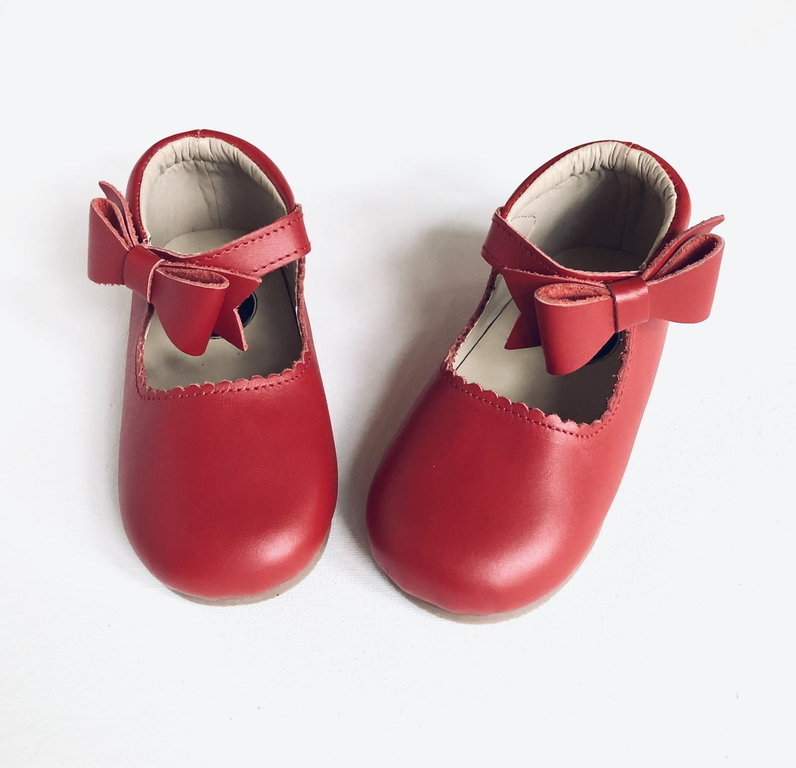 Red leather baby Mary Jane shoes with scalloped trim, strap, and decorative bow, displayed on white background.