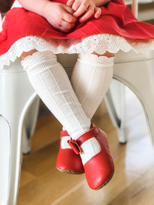 Toddler wearing red dress, lace-trimmed socks, and red Mary Janes with buckle strap, seated on white chair indoors.