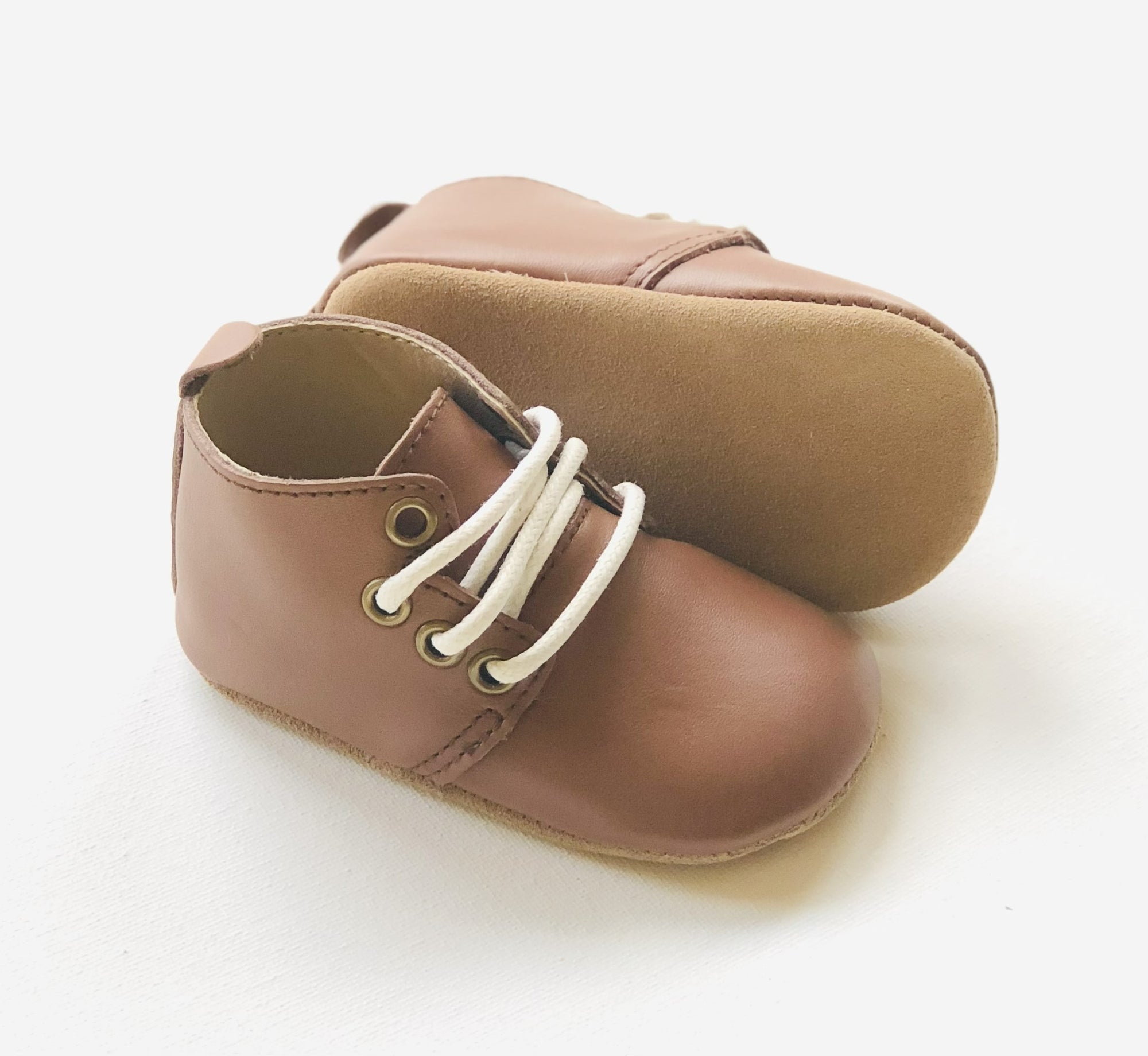 Brown leather baby shoes with white laces and soft suede sole, one upright and one on its side to show sole texture.