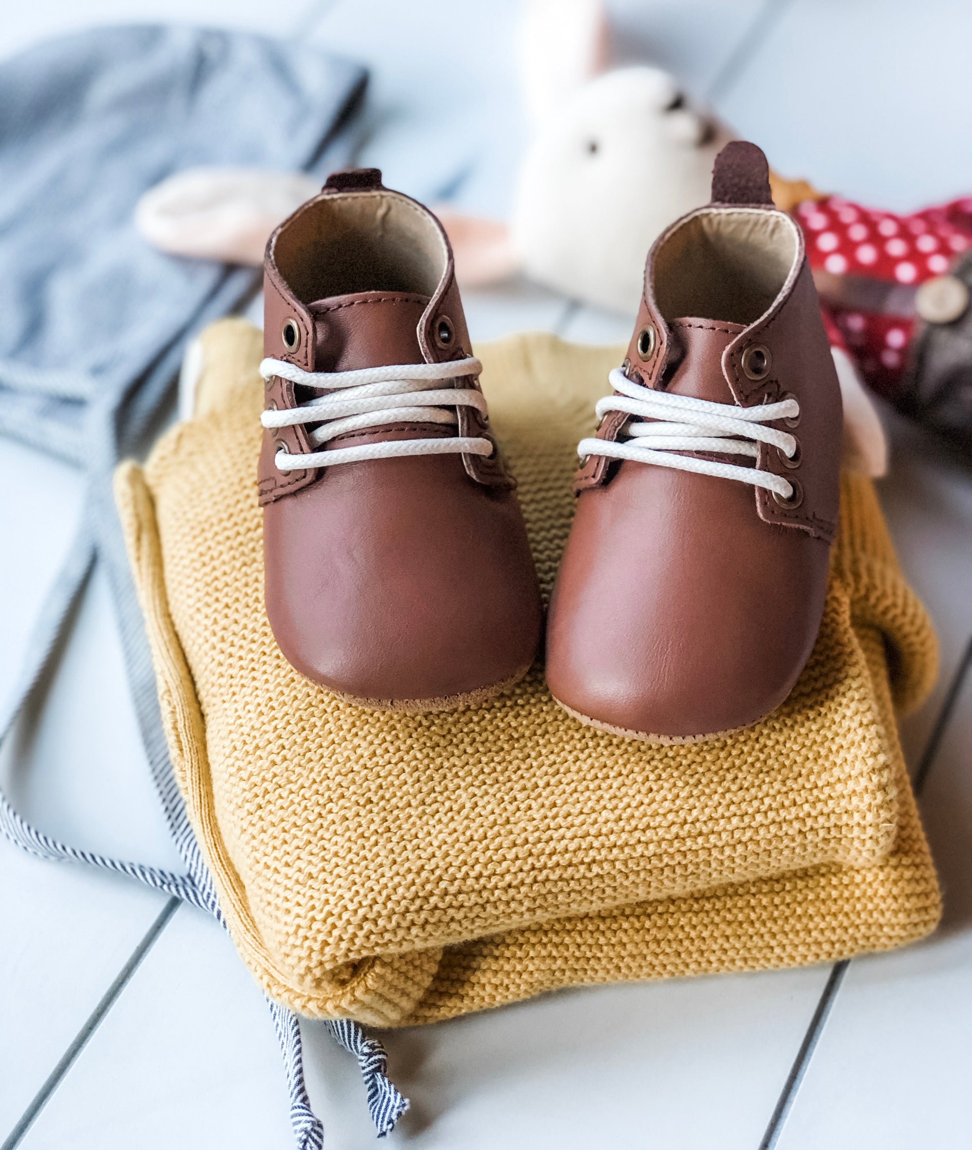 Brown leather baby Oxford shoes with white laces styled on folded yellow knit sweater beside plush bunny in red polka‑dot outfit.