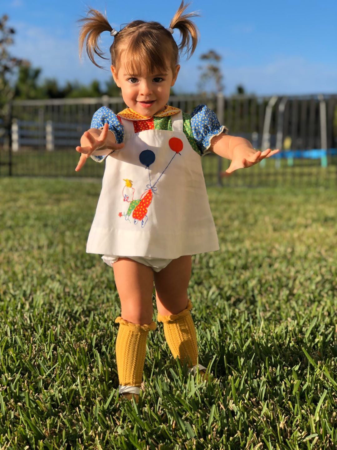 A young child with brown pigtails walks across a sunlit grassy yard, wearing a white dress with colorful balloon and animal designs, yellow knee-high socks, and bloomers—capturing playful movement, vintage charm, and outdoor joy.