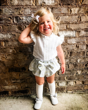 Smiling toddler with blonde hair and a white bow poses in front of a rustic brick wall, wearing a ruffled white top, bow-tied shorts, knee-high socks, and white shoes—blending vintage elegance with playful confidence.