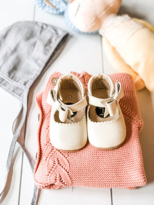 Flat lay of baby girl outfit with white shoes, pink knit sweater, striped bonnet, plush toys, and Peter Rabbit book – cozy and curated nursery styling.