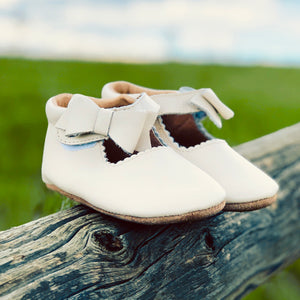 White Mary Jane baby shoes with scalloped trim and bows, styled on rustic wooden fence with green field background – outdoor lifestyle shot.
