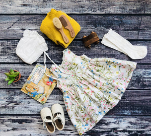 Flat lay of baby girl outfit with floral dress, white shoes, lace socks, bonnet, sweater, and Peter Rabbit book – vintage-inspired nursery styling.