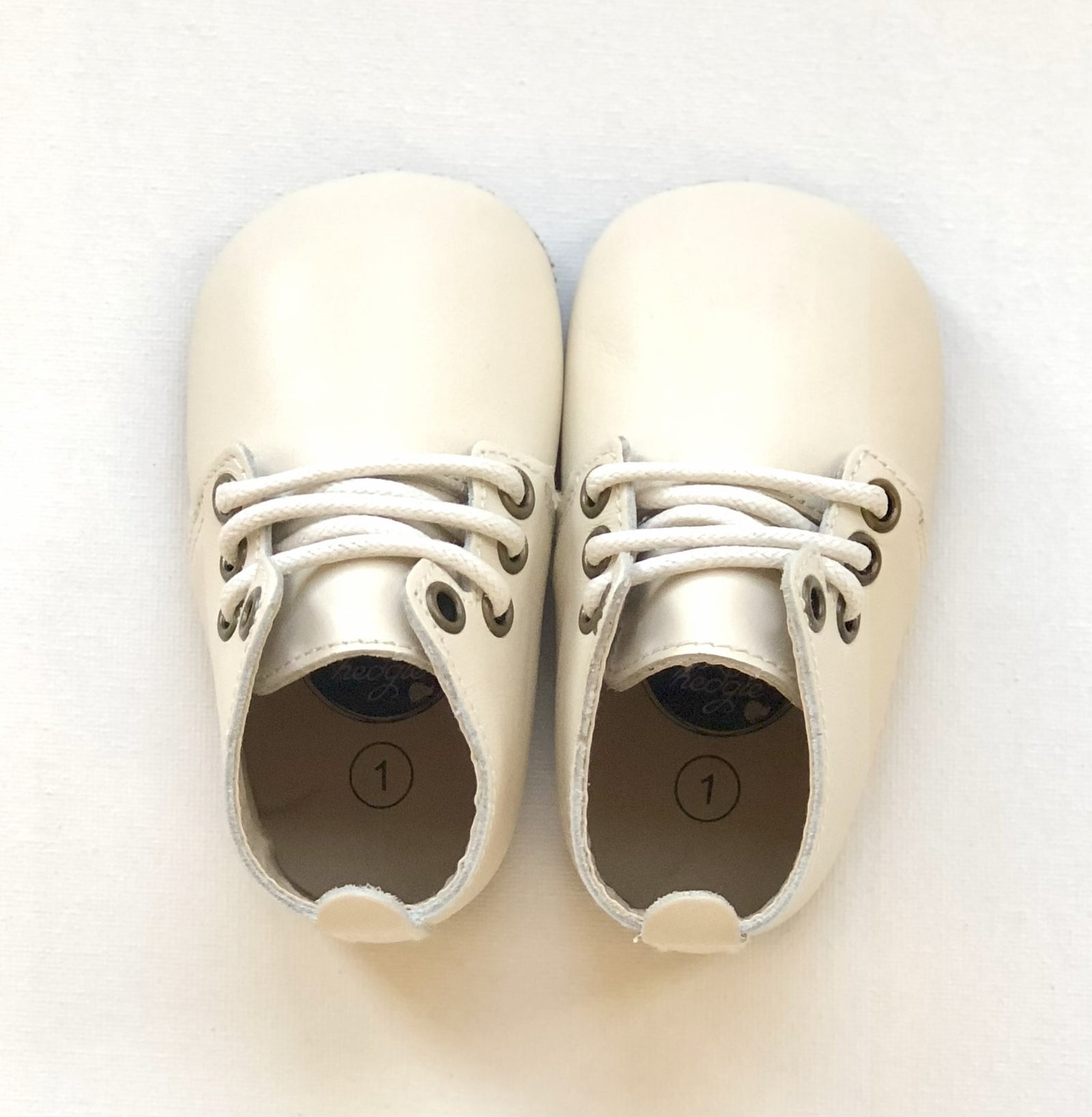 Cream leather baby shoes with white laces and size “1” insole marking, positioned side by side on light surface.