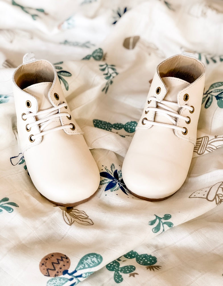 White leather baby Oxford shoes with gold eyelets and white laces, displayed on whimsical botanical‑print fabric background.
