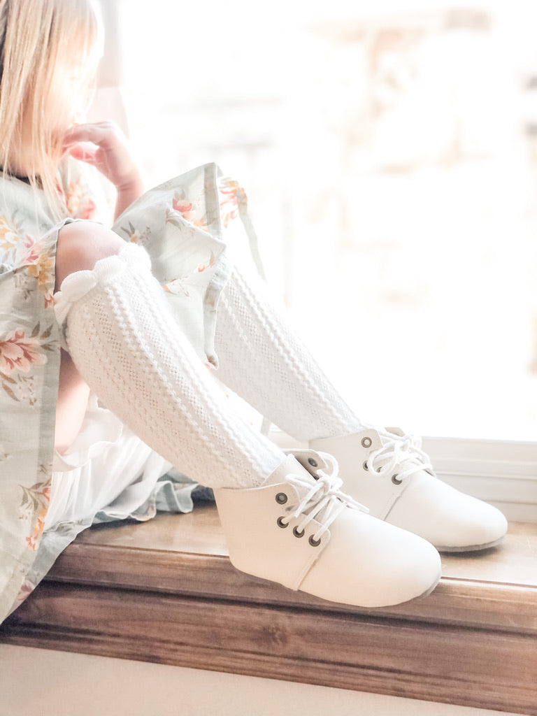 Toddler in floral dress and lace‑trimmed socks wearing white lace‑up ankle boots, sitting on wooden windowsill in natural light.