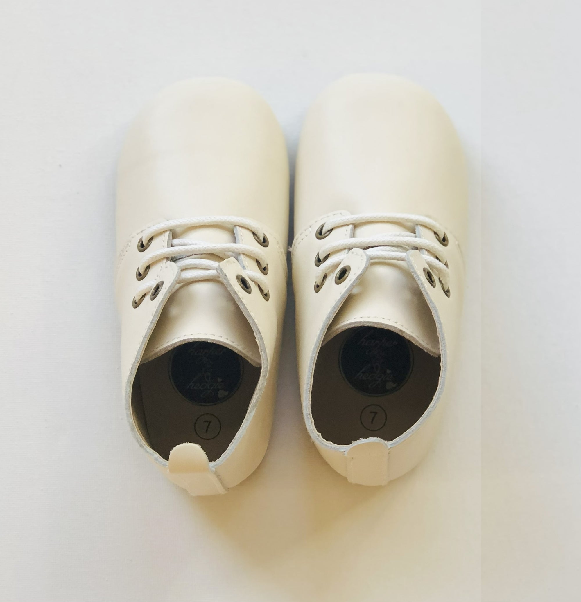 Cream leather baby Oxford shoes with white laces, pull tab, and size “7” insole branding, positioned side by side on white surface.