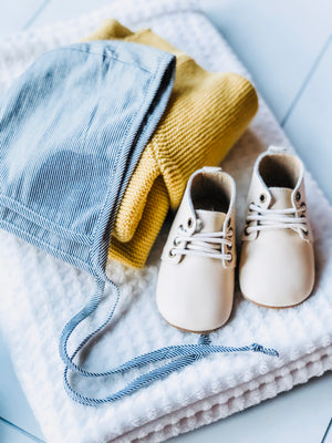 Cream leather baby shoes with white laces styled on white textured blanket alongside mustard knit sweater and blue‑striped bonnet.