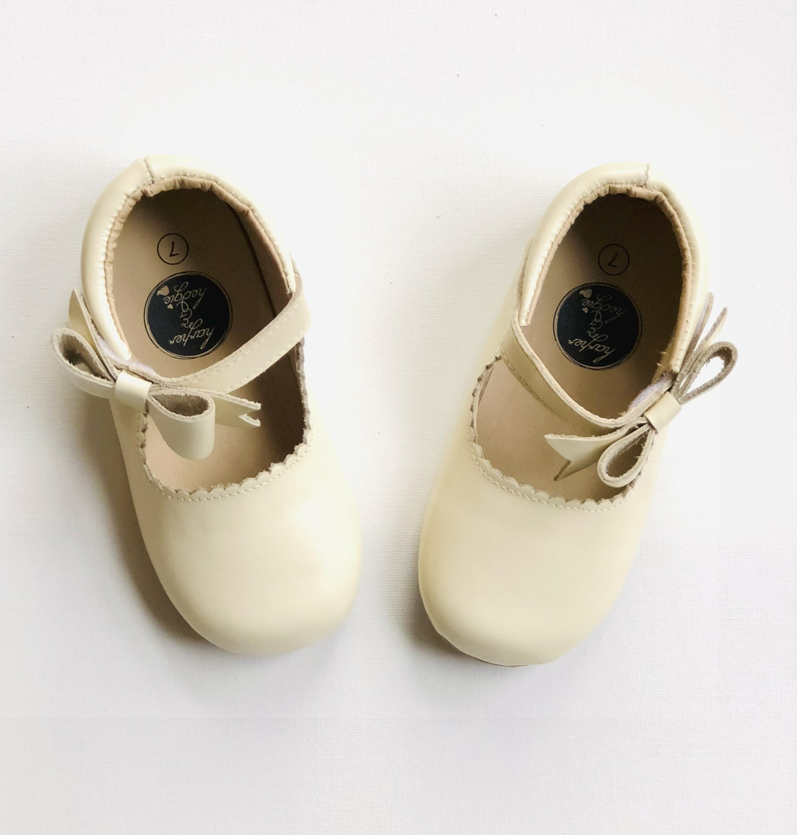 Cream-colored baby Mary Jane shoes with scalloped trim and front bows, styled for infants and toddlers.