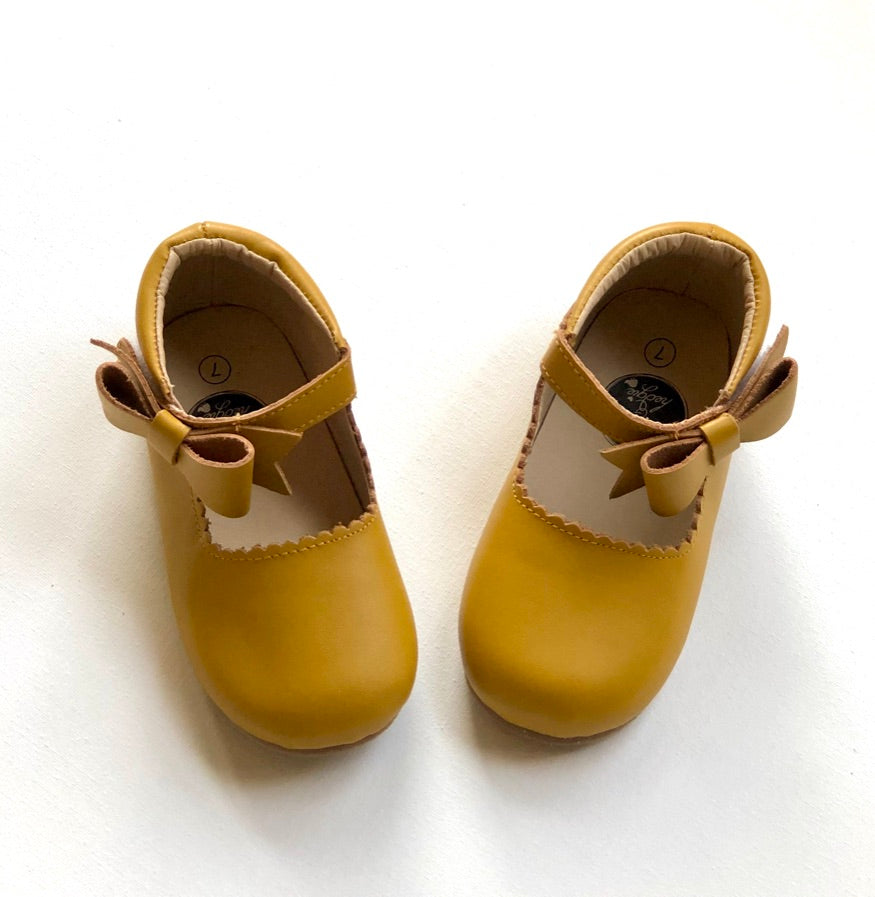 Mustard yellow Mary Jane baby shoes with scalloped trim and side bows, styled for toddlers and infants.