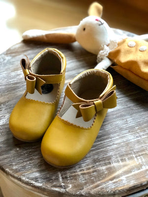 Mustard yellow baby shoes with brown bows and scalloped trim, styled beside a rabbit doll in matching outfit on wooden surface.