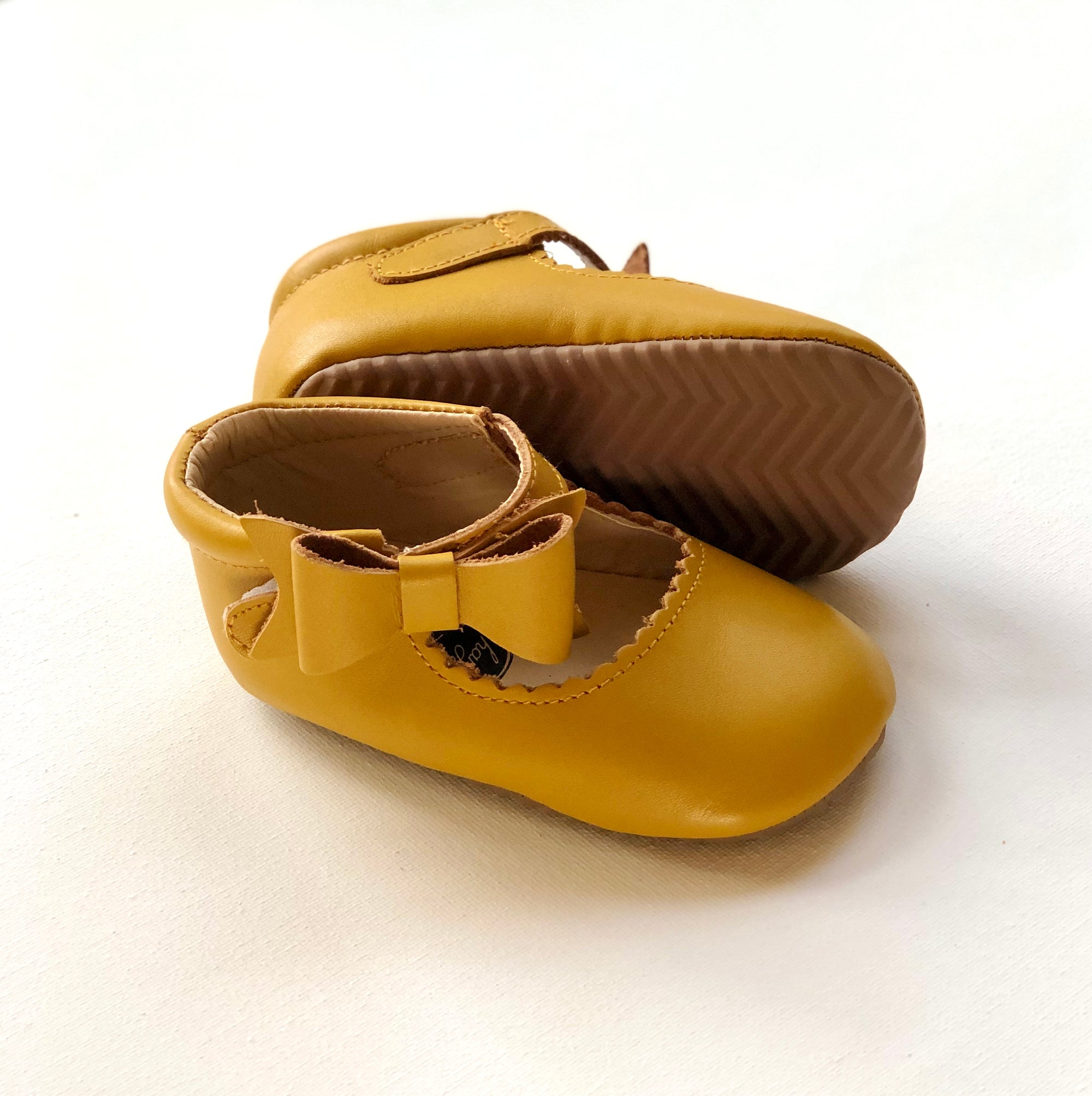 Mustard yellow baby Mary Jane shoes with front bows and zigzag tread sole, one upright and one on its side to show detail.