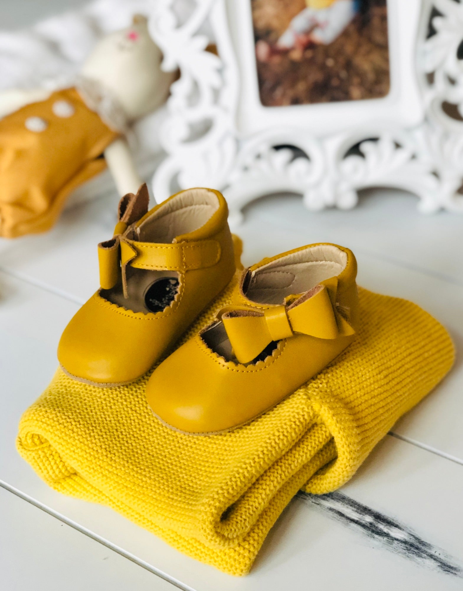 Mustard yellow baby Mary Jane shoes with bows, styled on folded knit sweater beside ornate picture frame and soft doll, creating a cozy nursery vibe.